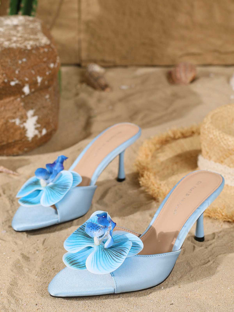 A pair of light blue satin mule heels with a pointed toe and a large, layered 3D fabric flower on the vamp, pictured in a beach-themed setting.