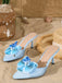 A pair of light blue satin mule heels with a pointed toe and a large, layered 3D fabric flower on the vamp, pictured in a beach-themed setting.