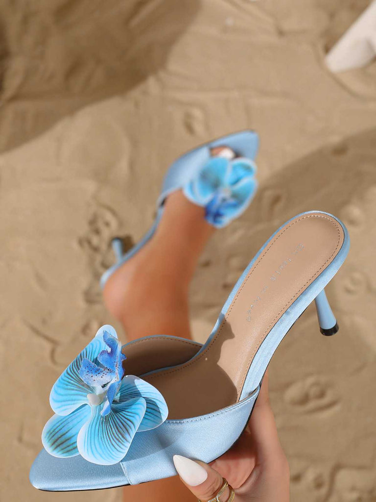 Angled shot of a single light blue satin mule showcasing the slender stiletto heel and floral embellishment.